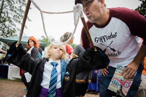 A child in a wizard costume plays with a bubble wand while an adult makes a funny face nearby at a festive event.