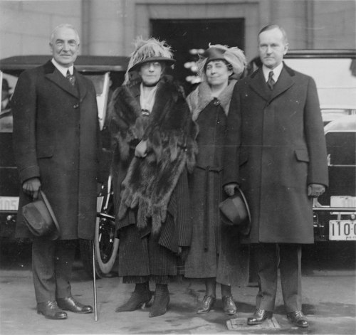 A black-and-white photo of four elegantly dressed individuals standing outside near a vintage car.
