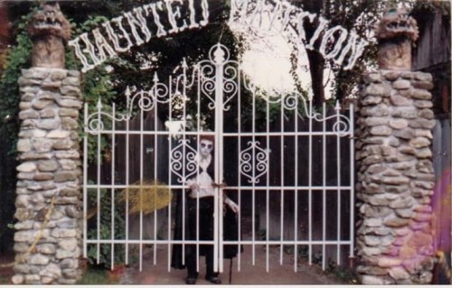A spooky gate labeled "Haunted Mansion" with a ghostly figure standing in front, surrounded by stone pillars and greenery.