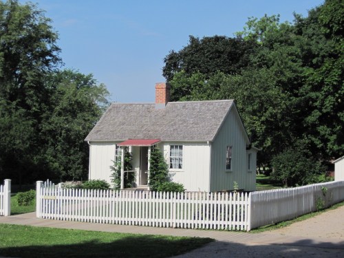 A small, light green house with a red roof, surrounded by trees and a white picket fence on a sunny day.