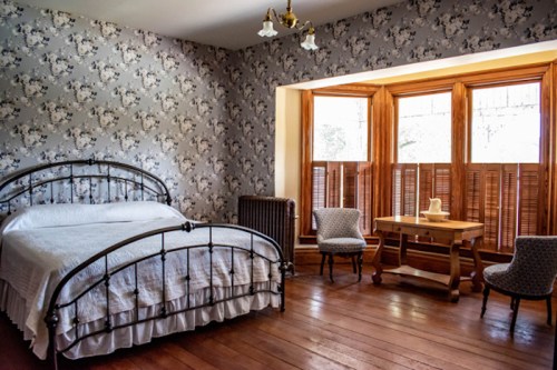 Cozy bedroom with floral wallpaper, a metal bed, wooden furniture, and large windows with wooden shutters.