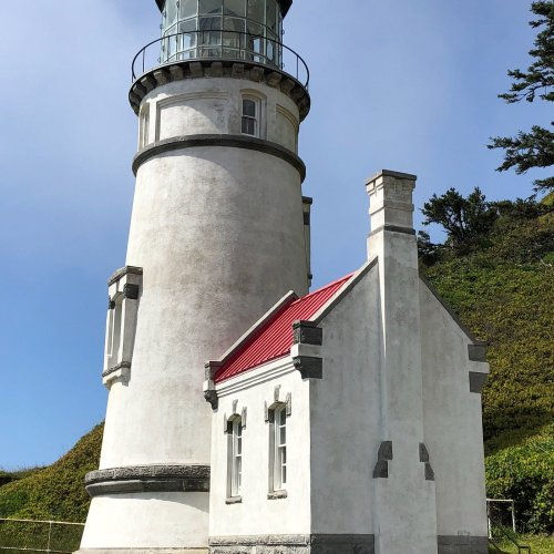 A tall lighthouse with a glass lantern room, surrounded by greenery and a clear blue sky.