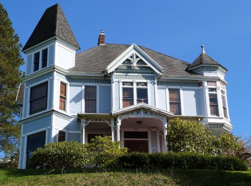 A large, Victorian-style house with a pointed tower, blue exterior, and lush greenery in front against a clear blue sky.