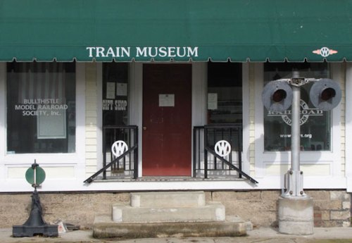 Exterior of a train museum with a green awning, featuring a sign, entrance door, and a railroad signal beside the steps.