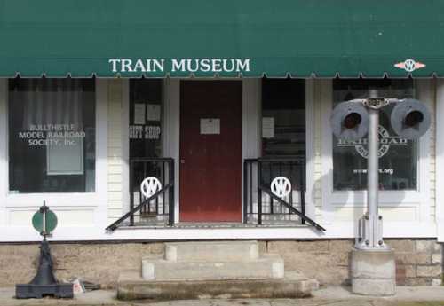 Exterior of a train museum with a green awning, featuring a sign, entrance door, and a railroad signal beside the steps.