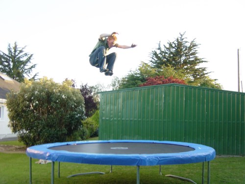 A person jumps high above a trampoline in a backyard, surrounded by trees and a green shed.