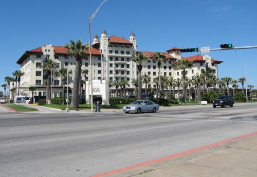 A large hotel with a red-tiled roof and palm trees, set against a clear blue sky, viewed from across the street.