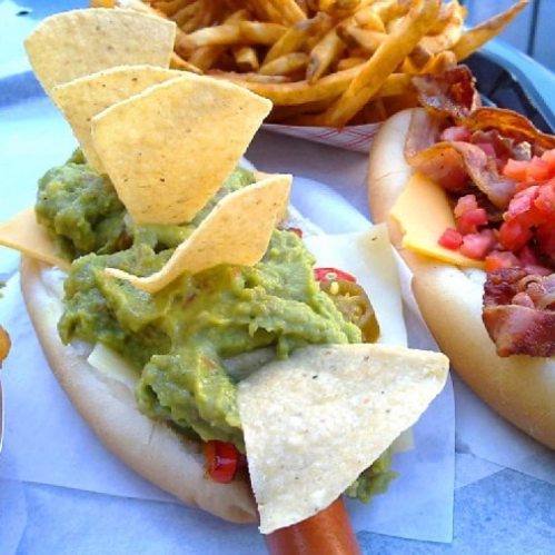 A hot dog topped with guacamole and tortilla chips, served alongside a basket of French fries.