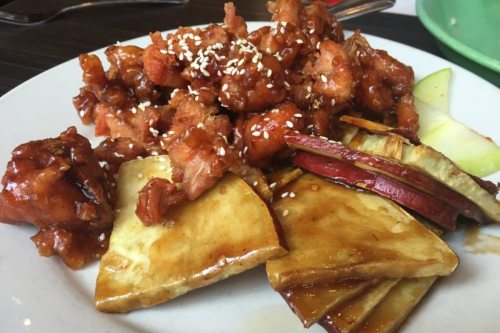 A plate of crispy orange chicken topped with sesame seeds, served with sliced apples and sweet potato.