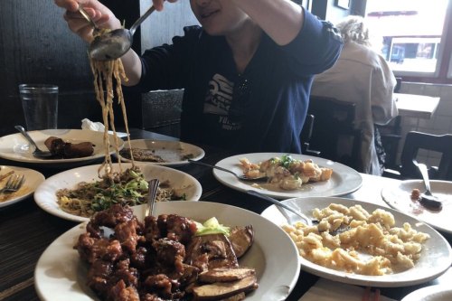 A person serves noodles from a bowl at a table filled with various dishes, including chicken and vegetables.