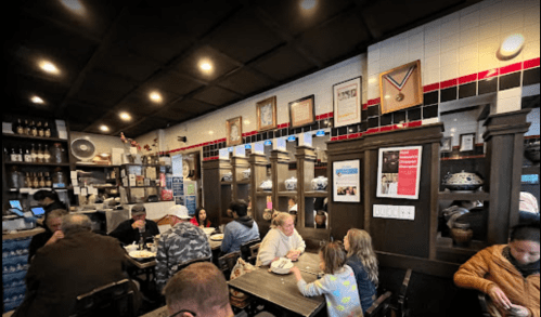 A cozy restaurant interior with diners at tables, shelves of dishes, and framed art on the walls.
