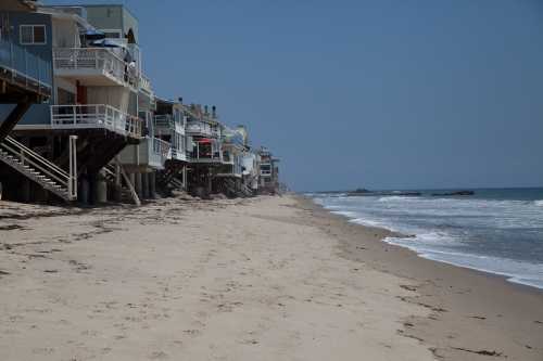 A sandy beach lined with beach houses on stilts, with gentle waves lapping at the shore under a clear blue sky.