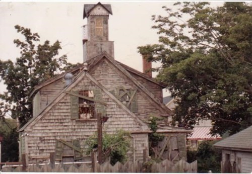 A dilapidated wooden house with boarded windows and a tall, weathered tower, surrounded by overgrown trees.