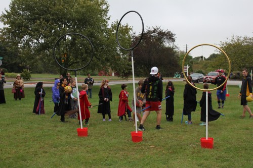 A group of children in costumes play a game with large hoops set up on a grassy field, surrounded by trees.