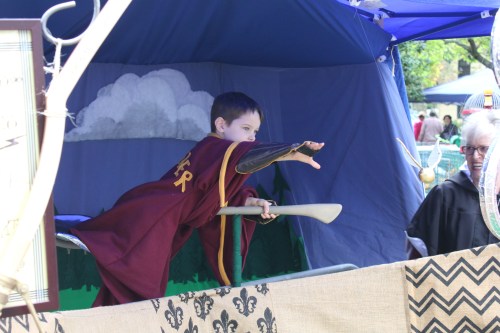 A young boy in a maroon costume points a toy sword from a decorated booth at an outdoor event.