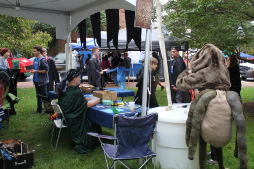 A lively outdoor event with people in costumes, booths, and tents, set in a grassy area.