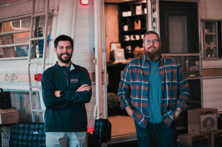 Two men stand in front of a vintage camper, smiling, with one wearing a sweater and the other in a plaid shirt.