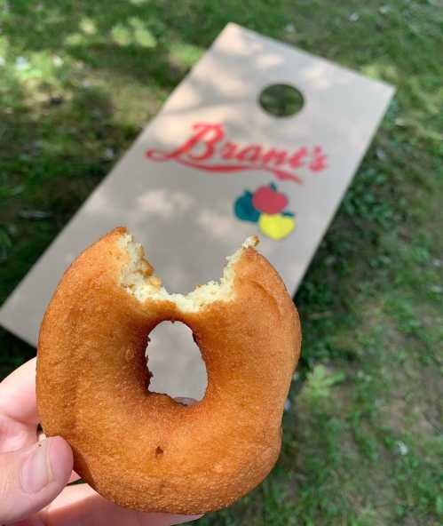 A hand holding a bitten donut with a hole, in front of a cornhole board with "Bran's" logo on it.