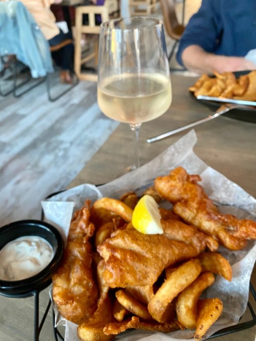 A basket of golden fried fish and chips with a lemon wedge, accompanied by a glass of white wine.
