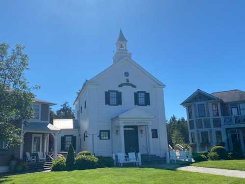 A white church with a steeple, surrounded by green grass and nearby houses under a clear blue sky.