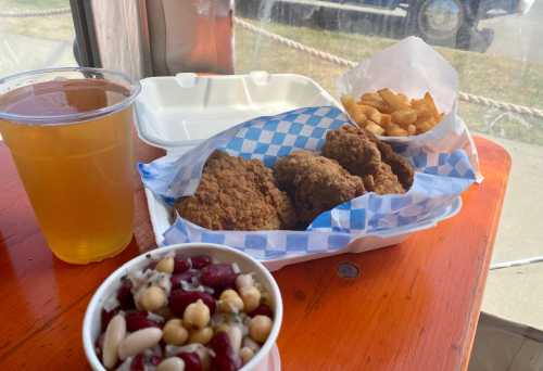 A takeout meal featuring fried chicken, fries, a bean salad, and a cup of beer on a wooden table.