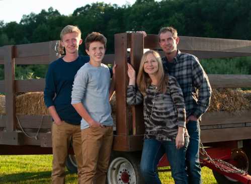 A family of four poses together by a wooden wagon in a sunny outdoor setting.