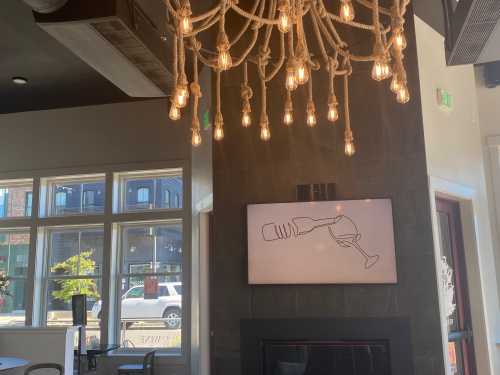 A cozy interior featuring a rope chandelier and a wall art of a wine glass, with large windows letting in natural light.