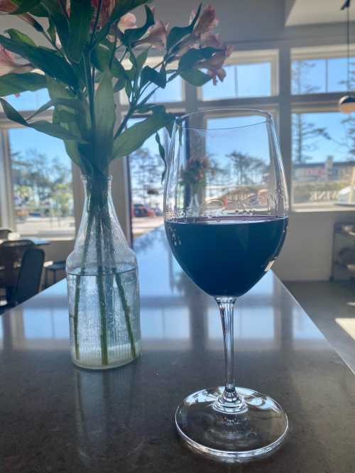 A glass of red wine sits on a table next to a vase of flowers, with a bright, sunny background.