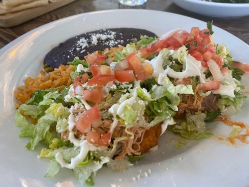 A plate of enchiladas topped with lettuce, tomatoes, sour cream, and cheese, served with rice and black beans.