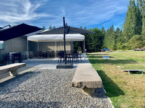 Outdoor seating area with a tent, tables, and chairs, surrounded by grass and trees under a blue sky.