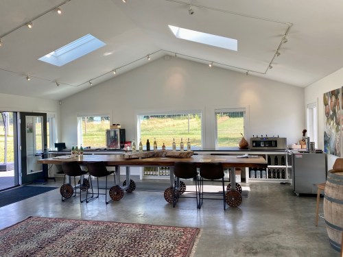 A modern tasting room with a long wooden bar, high stools, and large windows overlooking a green landscape.