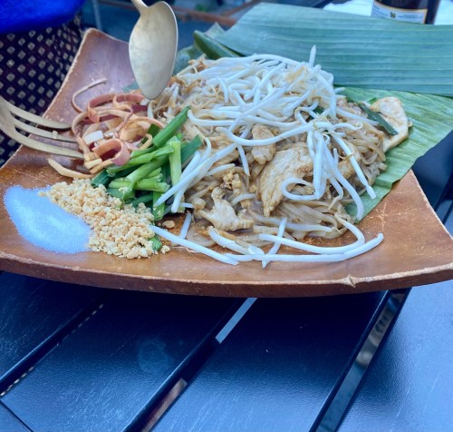 A wooden plate with pad thai, garnished with bean sprouts, green onions, peanuts, and lime on a table.