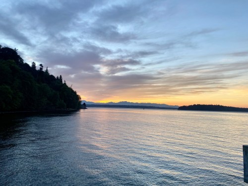 A serene sunset over calm waters, with silhouettes of trees and mountains in the background.