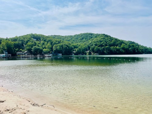 A serene lake surrounded by lush green hills under a clear blue sky, with a sandy shore in the foreground.