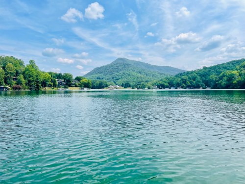 A serene lake surrounded by lush green trees and a distant mountain under a blue sky with fluffy clouds.