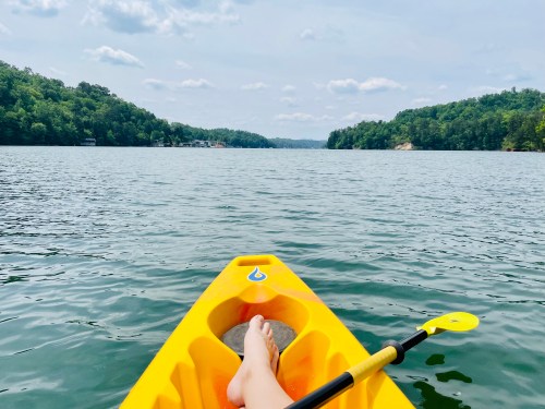 A person’s foot rests on the edge of a yellow kayak, with calm water and green hills in the background.