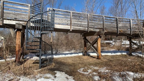A wooden walkway with a spiral staircase, surrounded by bare trees and patches of snow on the ground.