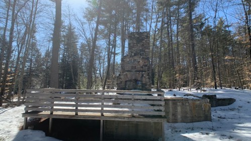 A stone chimney stands in a snowy forest, with a wooden bench in the foreground and trees surrounding the area.