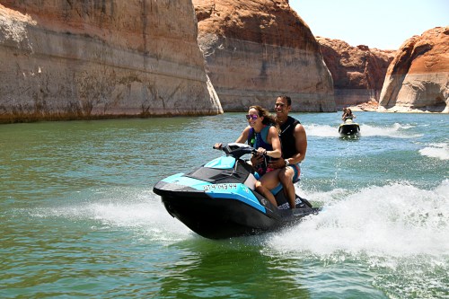 Two people ride a jet ski on a lake, surrounded by rocky cliffs and clear blue water, with another jet ski in the background.