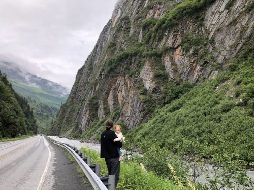 A person holds a child while standing by a scenic mountain road, surrounded by lush greenery and steep cliffs.