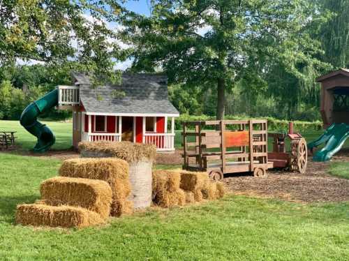 A colorful playground featuring a small playhouse, slides, and hay bales in a grassy area surrounded by trees.