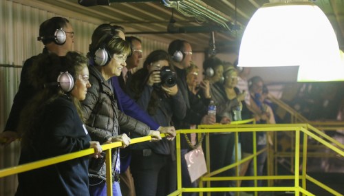 A group of people wearing headphones observes from a balcony, with some taking photos under bright lighting.