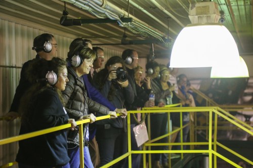 A group of people wearing headphones observes from a balcony, with cameras ready, under bright overhead lights.