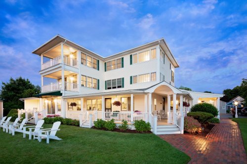 A charming three-story house with a wraparound porch, surrounded by green grass and white adirondack chairs at dusk.