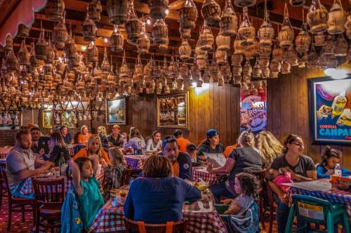 A lively restaurant interior with families dining at tables, decorated with hanging lanterns and checkered tablecloths.