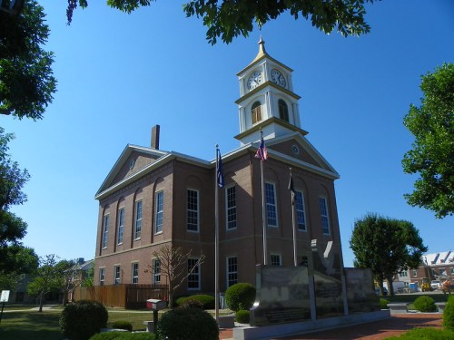 Historic brick building with a clock tower, surrounded by trees and flags, under a clear blue sky.
