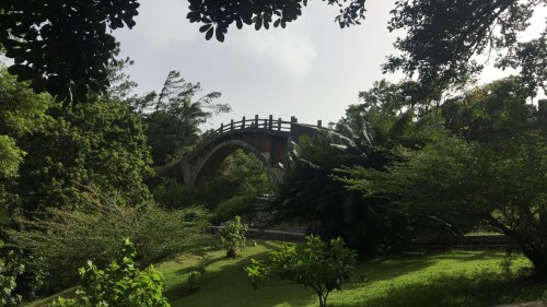 A scenic view of a curved bridge surrounded by lush greenery and trees under a cloudy sky.