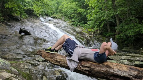 A person relaxes on a log by a flowing stream, surrounded by lush greenery and rocky terrain.