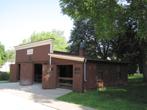 A rustic brown building with open doors, surrounded by green trees and grass, resembling a historic barn or workshop.