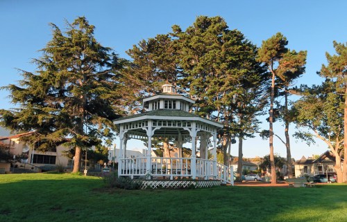 A white gazebo surrounded by tall trees in a sunny park setting.